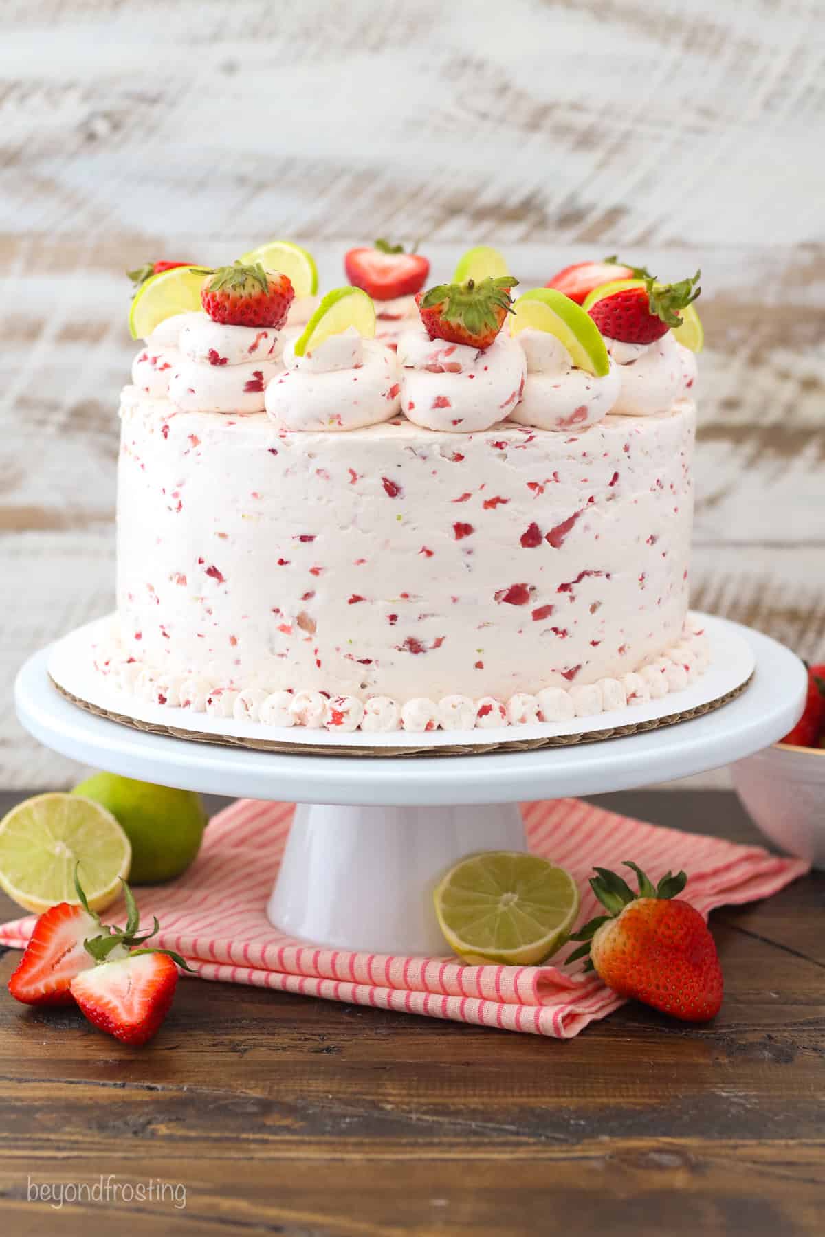 This homemade strawberry margarita cake has layers of lime and tequila-infused cake frosted with fresh strawberry Swiss meringue buttercream. A three-tier strawberry margarita layer cake frosted with strawberry buttercream and decorated with rosettes, fresh berries, and lime wedges on a cake stand.