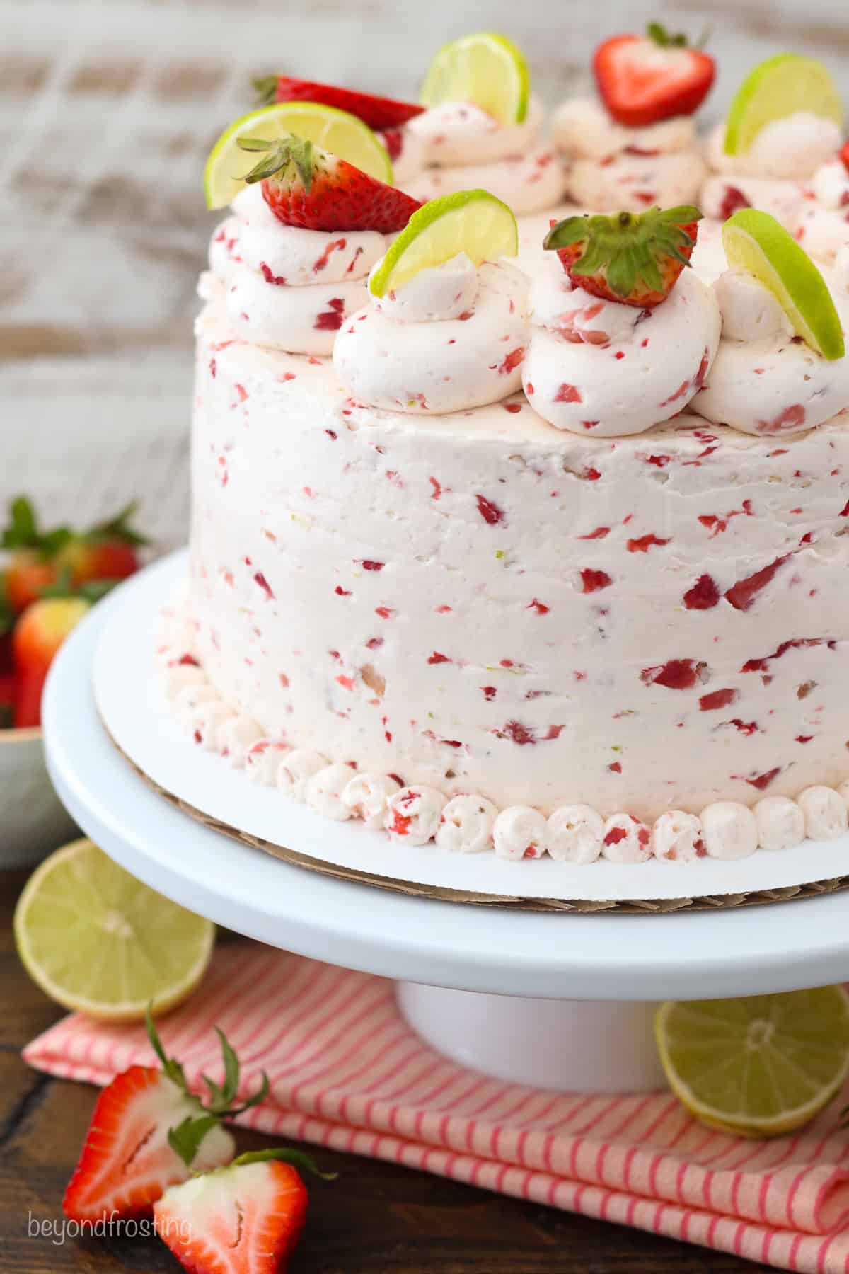 Close up of a three-tier strawberry margarita layer cake frosted with strawberry buttercream and decorated with rosettes, fresh berries, and lime wedges on a cake stand.
