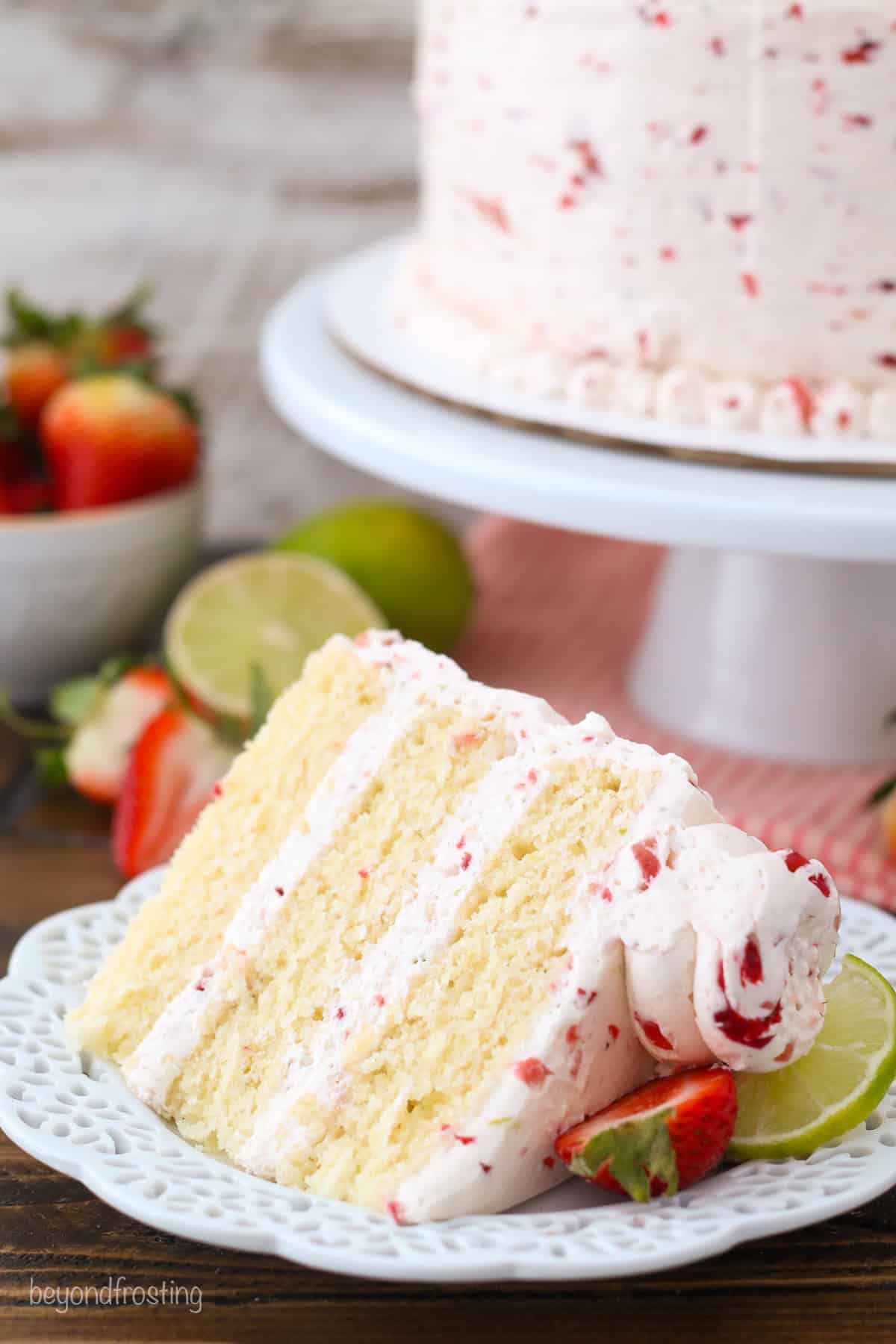 A slice of strawberry margarita layer cake on a plate with the rest of the cake on a cake stand in the background.