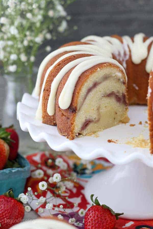 A Strawberry Swirl Bundt Cake with a slice taken out of it on a white cake plate with scallop edges