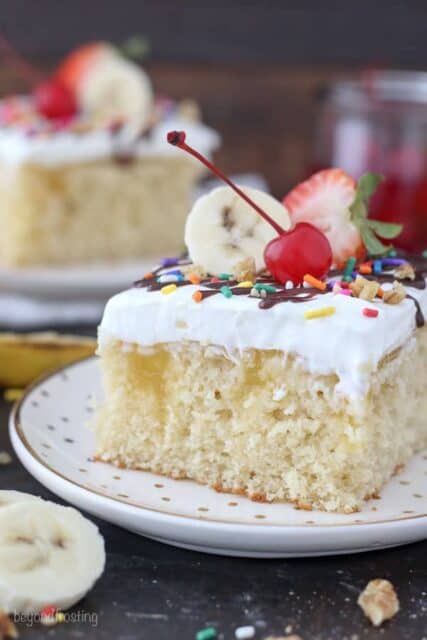 A slice of banana split cake on a gold polka dot plate. The vanilla cake is filled with a banana pudding and topped with sundae toppings