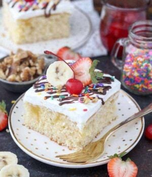 A beautifully decorated slice of cake garnished with a cherry, a sliced banana, a strawberry, sprinkles and hot fudge sauce.