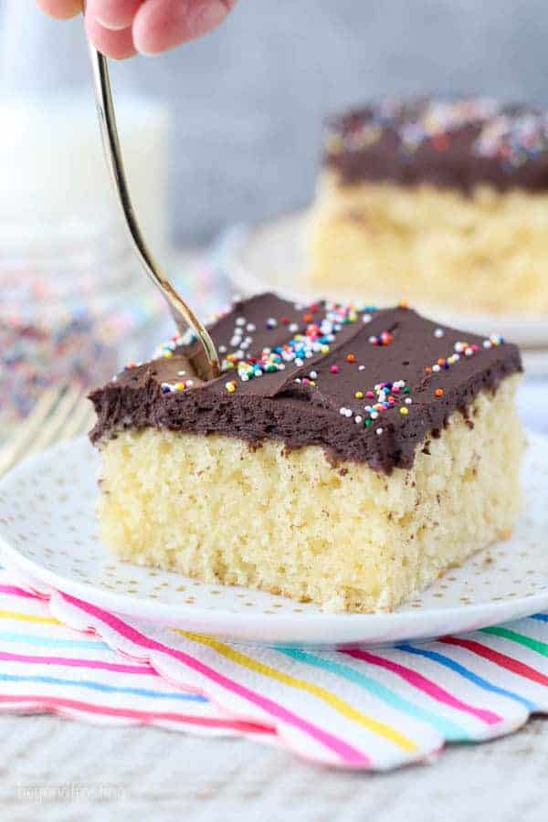 A gold fork sinking into a slice of yellow cake with chocolate frosting and sprinkles