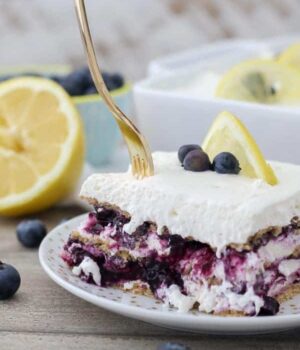 A gold fork digging into a layered icebox cake with blueberries and a lemon wedge on top.