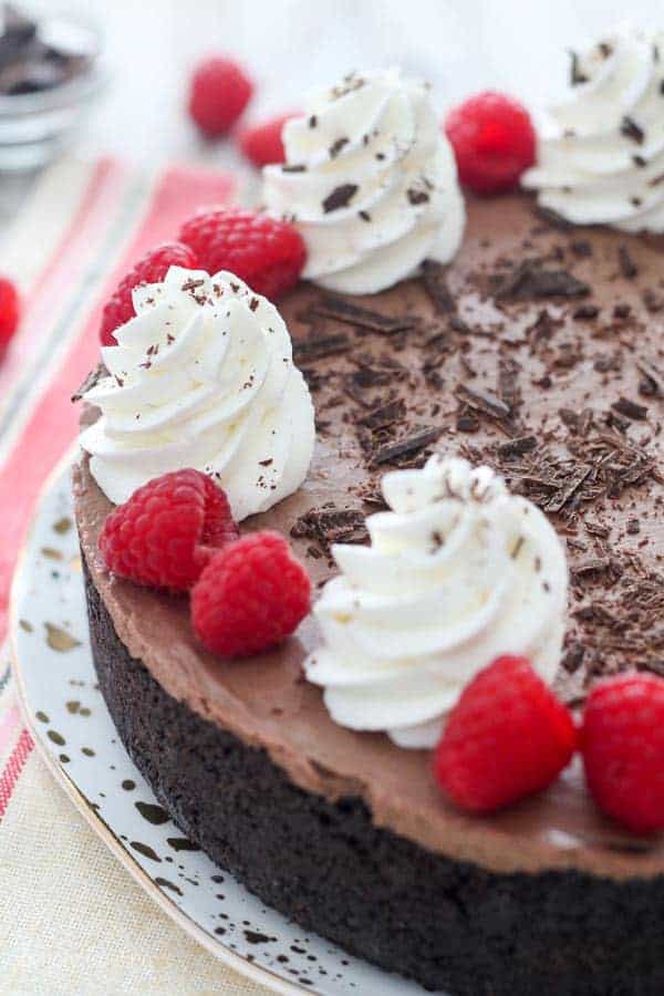 A large chocolate cheesecake garnished with whipped cream chopped chocolate and raspberries.