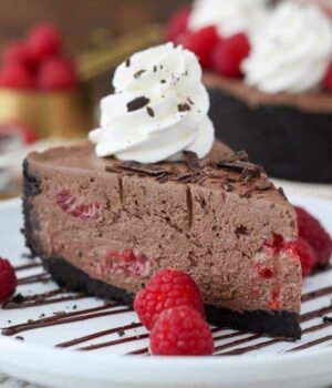 A big slice of chocolate cheesecake on a white plate drizzled with chocolate and garnished with raspberries