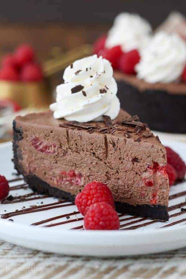 A big slice of chocolate cheesecake on a white plate drizzled with chocolate and garnished with raspberries