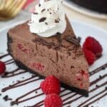A big slice of chocolate cheesecake on a white plate drizzled with chocolate and garnished with raspberries