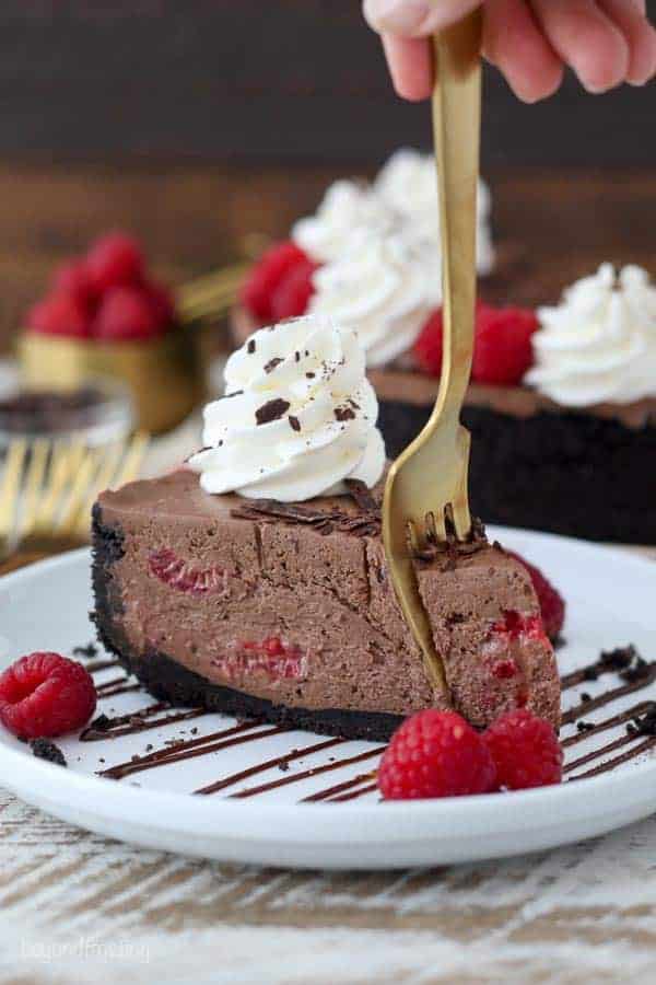 A gold fork sinking into a slice of chocolate cheesecake that is loaded with raspberries