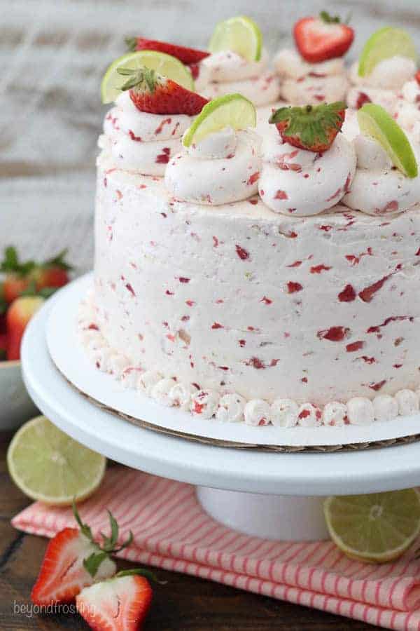 A close up of a strawberry and lime layer cake. The cake is sitting on a white cake stand with a pink striped napkin