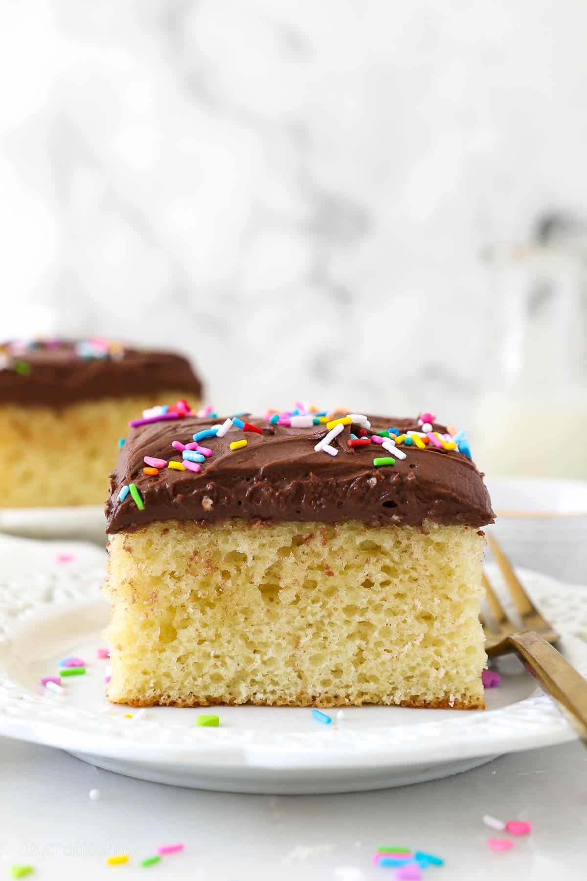 A slice of yellow cake with chocolate frosting and sprinkles on a white plate.