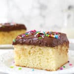 A slice of yellow cake with chocolate frosting and sprinkles on a white plate.