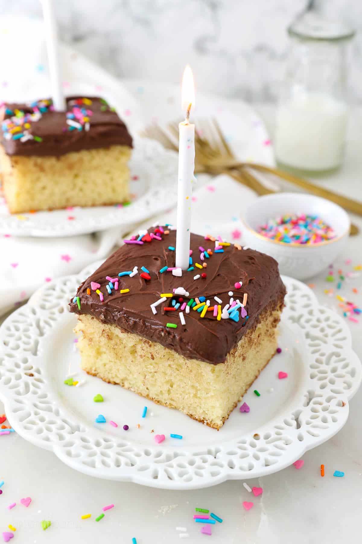 A slice of yellow cake with chocolate frosting and sprinkles on a white plate topped with a birthday candle.