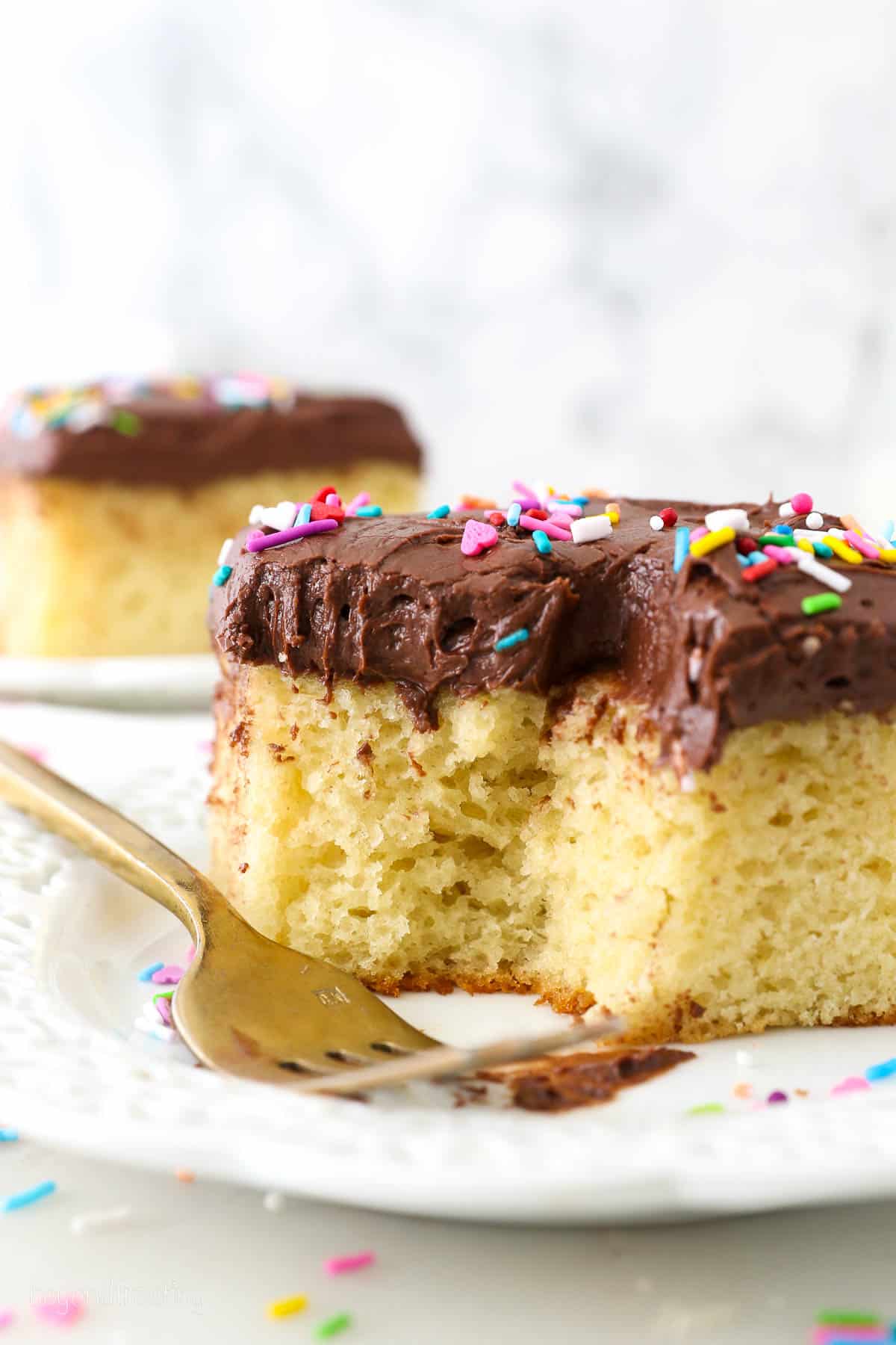 A slice of yellow cake with chocolate frosting and sprinkles on a white plate with a forkful missing.