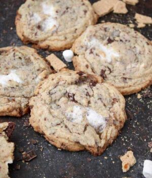 A bunch of cookies with melted marshmallow centers are laid out on a cookie sheet with a lot of crumbs and mini marshmallows