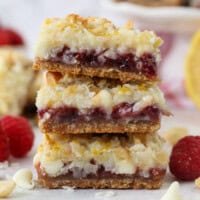 A stack of three raspberry lemon magic bars on a countertop next to raspberries and white chocolate chips.