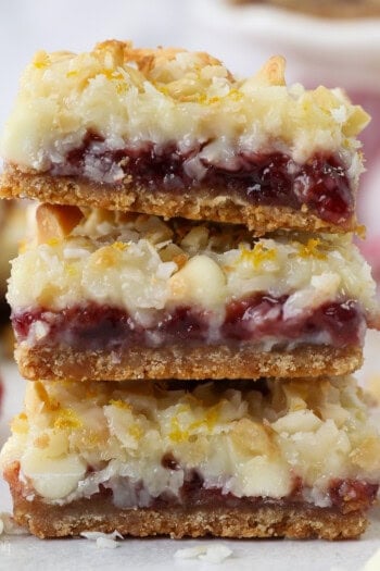 A stack of three raspberry lemon magic bars on a countertop next to raspberries and white chocolate chips.