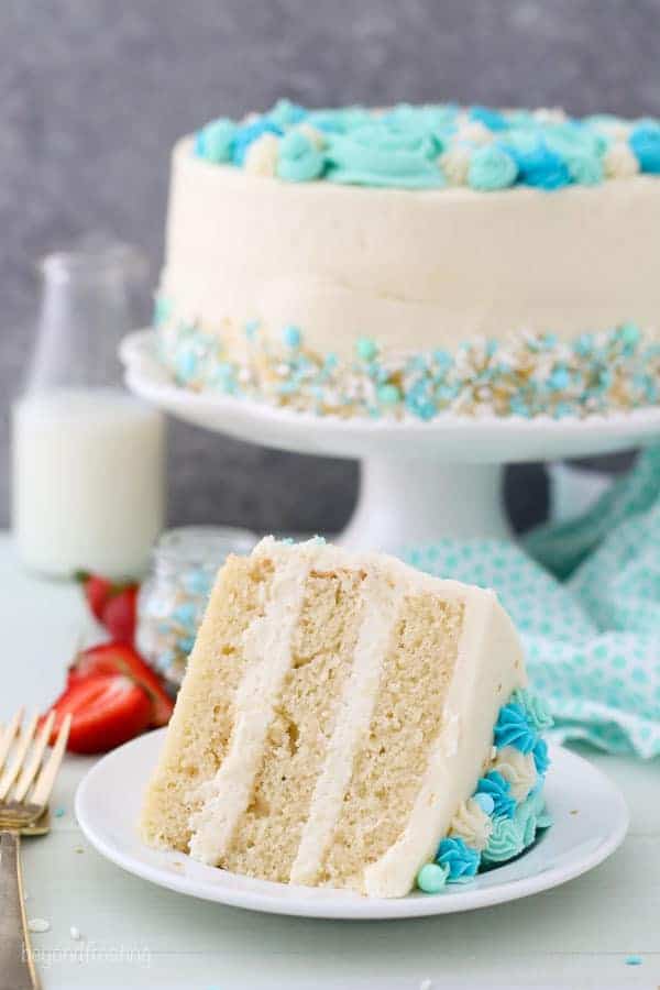 A Slice of Vanilla Layer Cake on a Plate Next to the Full Cake on a Cake Stand