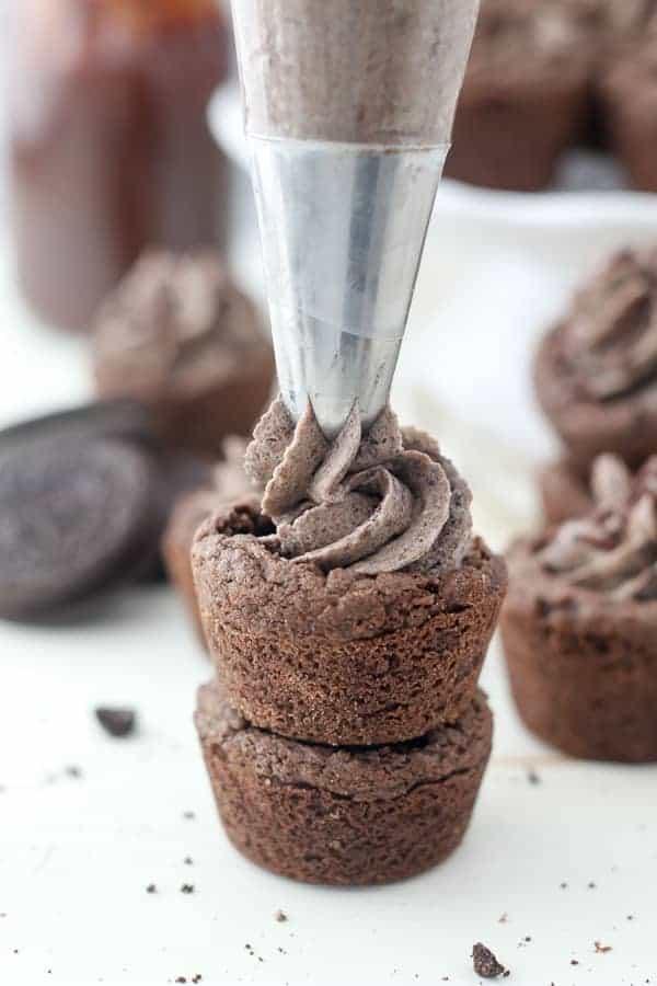 Two chocolate cookie cups stacked on top of one another. The one on top is being filled with an oreo filling from a piping bag