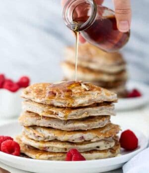 A small jar of maple syrup being poured over a stack of pancakes.