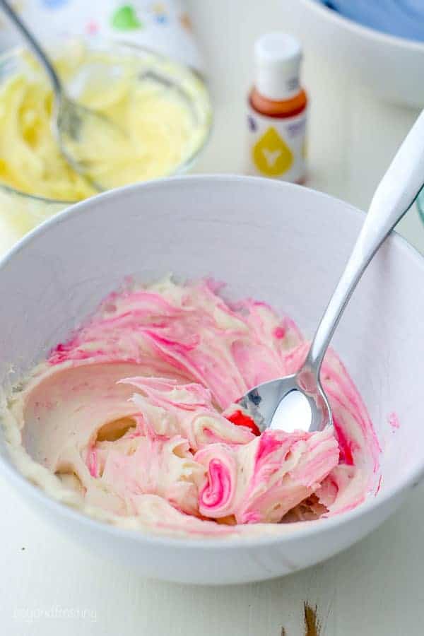 A white bowl filled with vanilla frosting that is being dyed pink. The pink color is swirled into the frosting