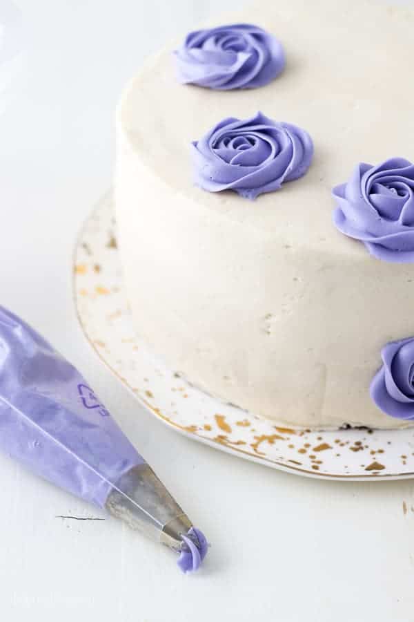 An icing bag filled with purple frosting is laying next to a cake decorated with several buttercream roses