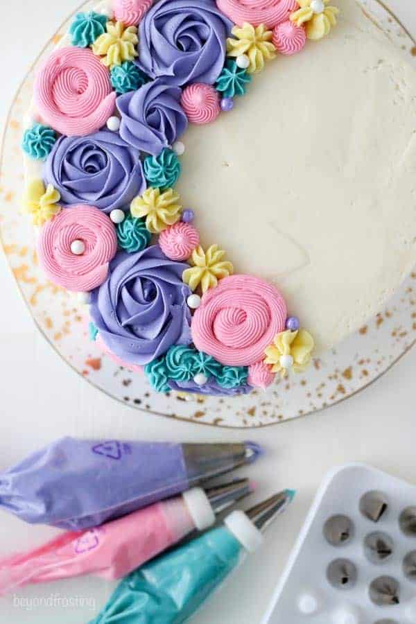 A close up overhead view of a layer cake decorated with purple buttercream roses and multicolor buttercream flowers