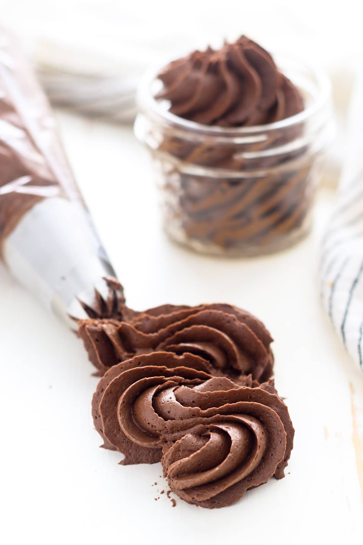 A piping tip pipes a squiggle of chocolate fudge frosting onto a white surface, with a jar of piped frosting in the background.