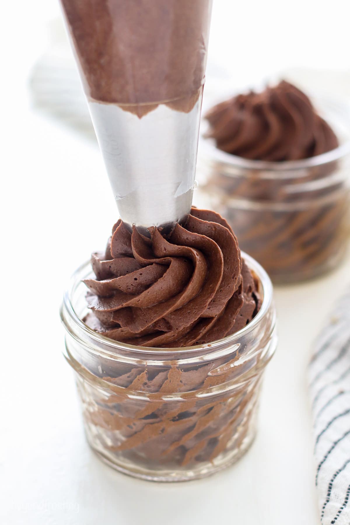 A piping tip pipes a swirl of chocolate fudge frosting into a glass jar, with a second jar of piped frosting in the background.