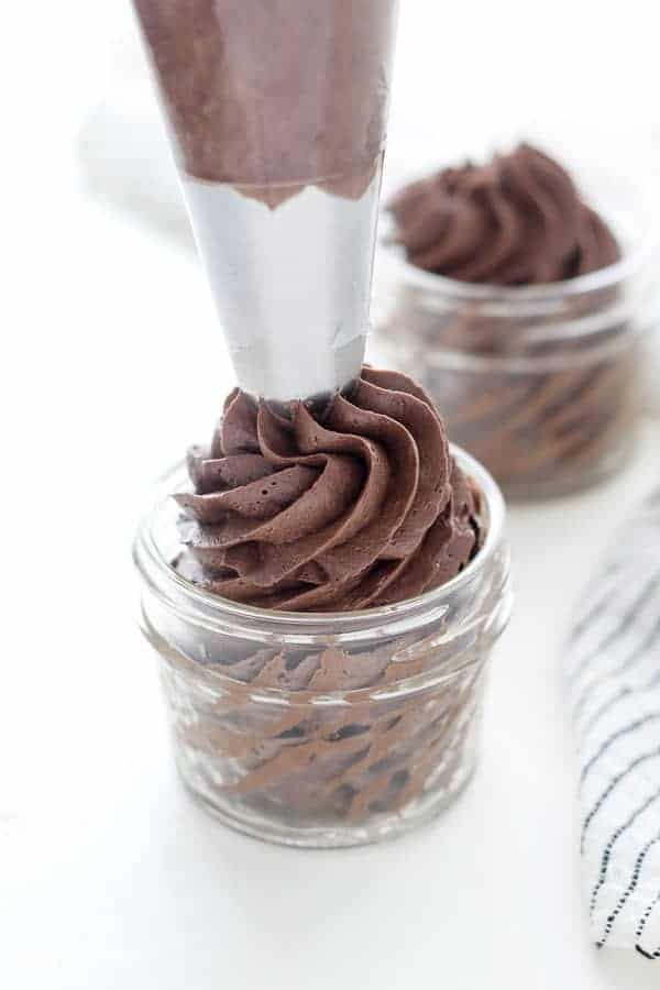 A large piping bag filling a small glass jar with silky chocolate buttercream frosting