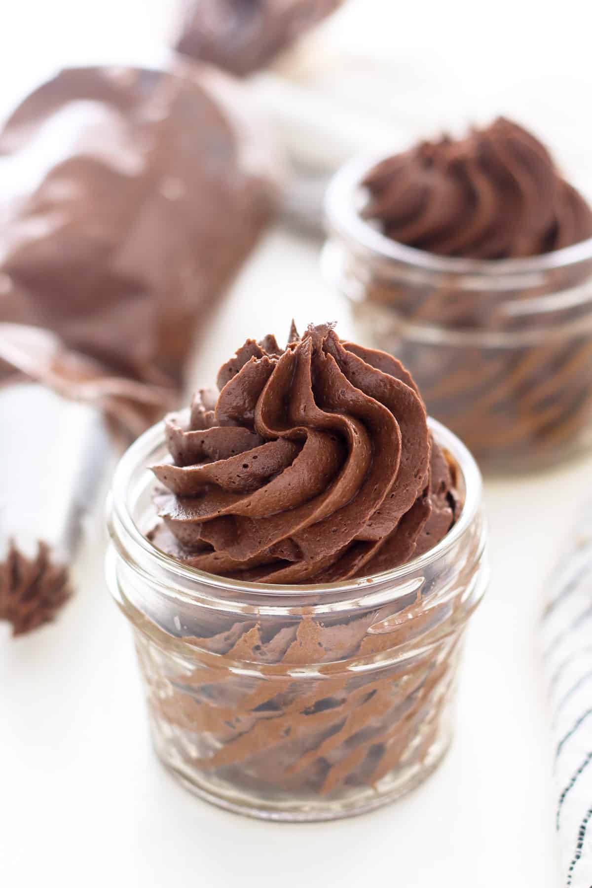 Chocolate fudge frosting swirled in a glass jar, with a second jar of frosting and a piping bag in the background.