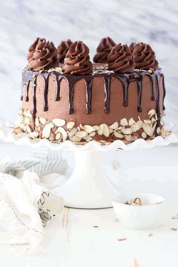 A gorgeous chocolate frosted cake with sliced almonds and a chocolate ganache drizzle on a white scalloped cake plate