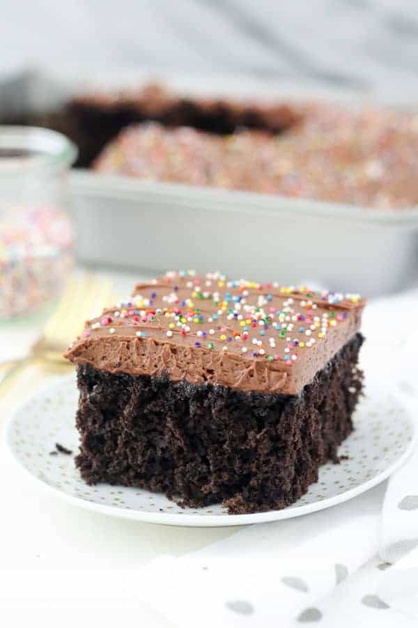 A beautiful slice of chocolate cake on a white plate. The cake is frosted with a chocolate frosting and sprinkles.