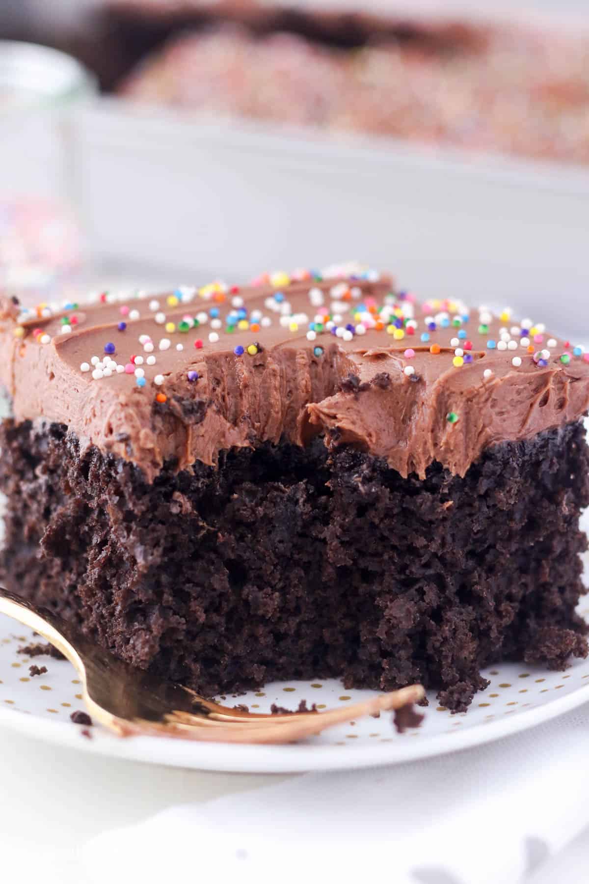 A slice of frosted chocolate cake topped with rainbow sprinkles on a white plate, with a forkful missing.