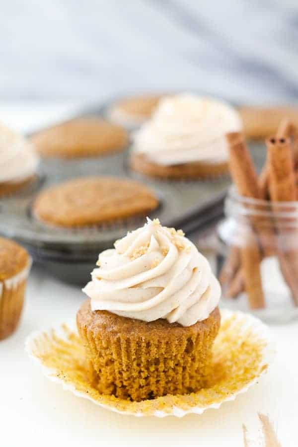 A pumpkin cupcake with the wrapper pulled away, topped with a swirl of cream cheese frosting
