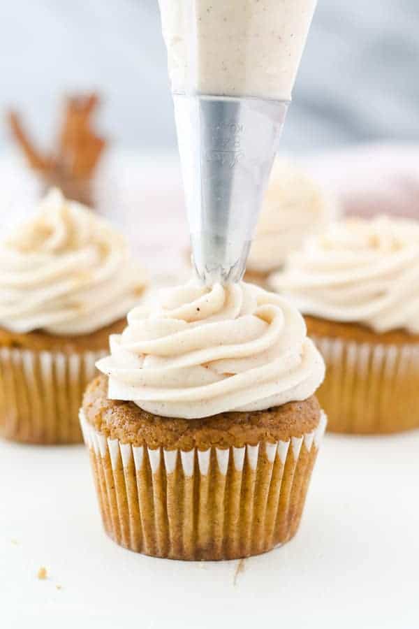 A pumpkin cupcake is piping a swirl of cream cheese frosting on top
