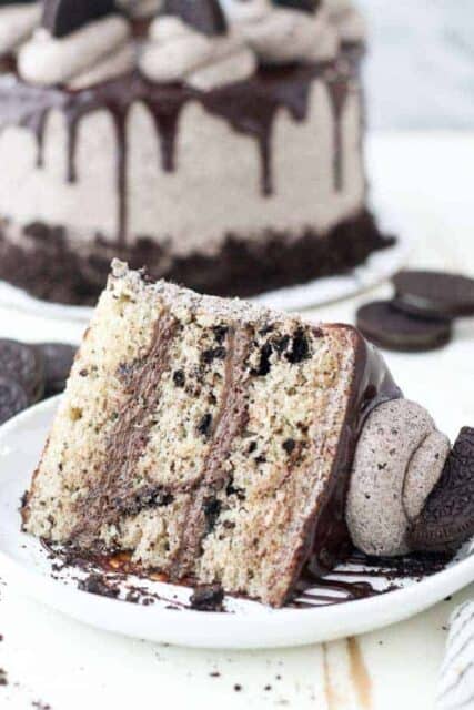 et gigantisk stykke kake på en hvit rimmet plate. Kaken er lastet Med knuste Oreos og Det er En stor porsjon Oreo frosting på toppen's a big dollop of Oreo frosting on top