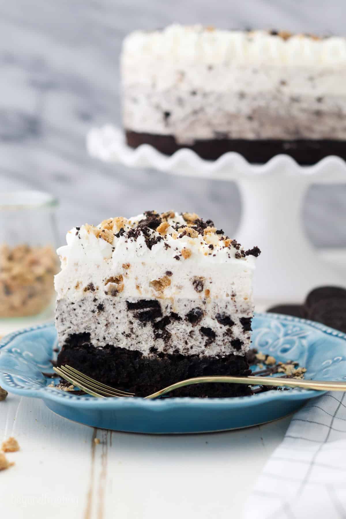 A slice of Oreo brookie mousse cake on a blue plate with a fork, and the rest of the cake on a cake stand in the background.
