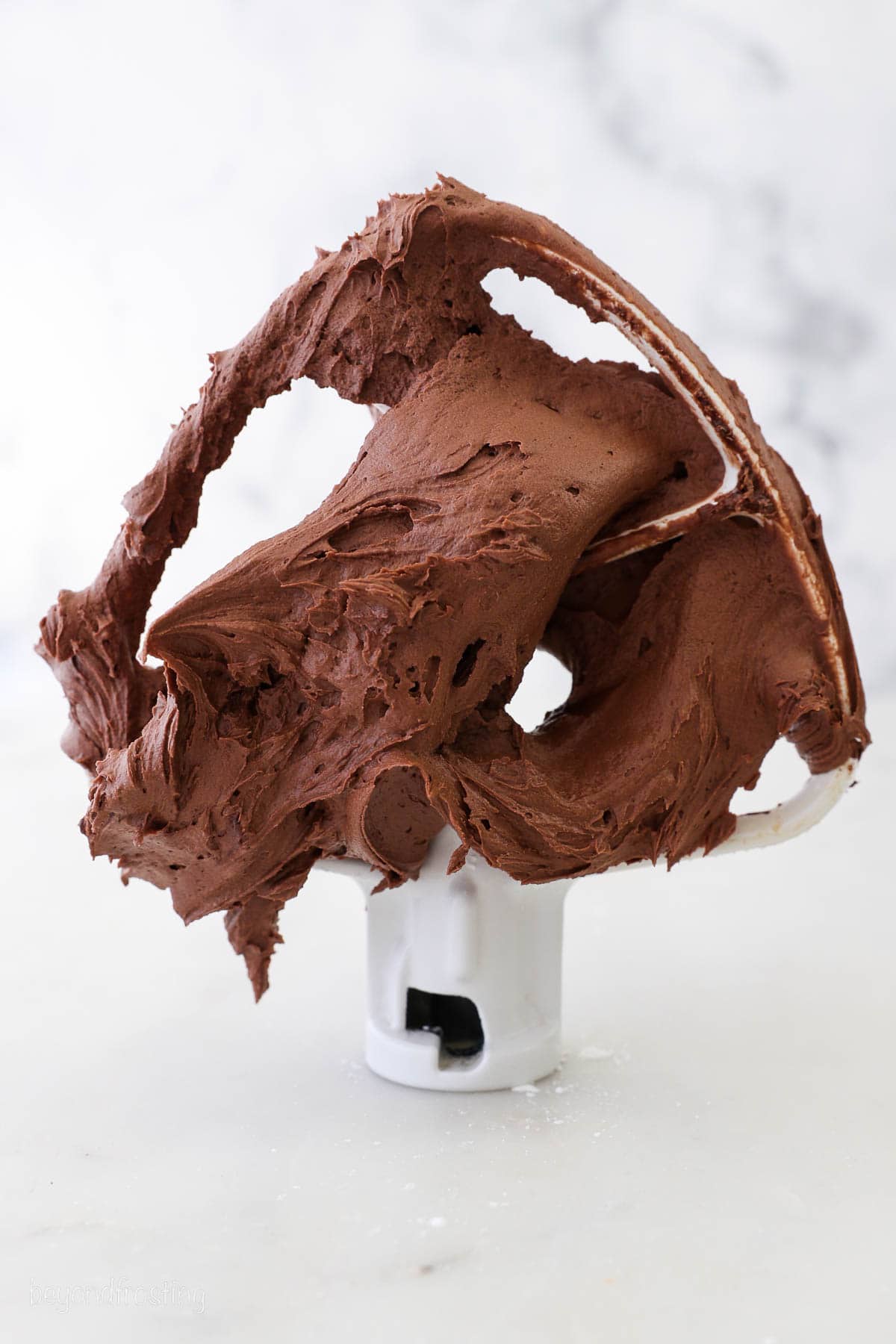 A stand mixer attachment propped upright on a countertop coated in thick chocolate cream cheese frosting.