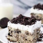A slice of cookies and cream cake on a white plate drizzled with hot fudge sauce and crushed Oreos