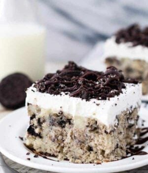 A slice of cookies and cream cake on a white plate drizzled with hot fudge sauce and crushed Oreos