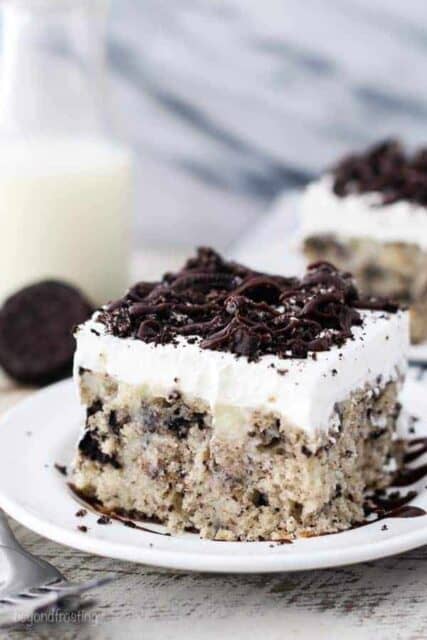 A slice of cookies and cream cake on a white plate drizzled with hot fudge sauce and crushed Oreos