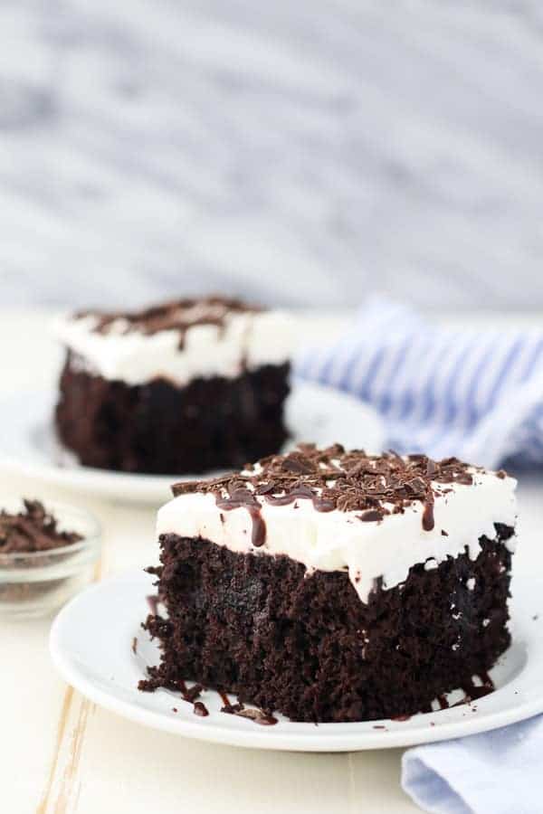 A slice of chocolate poke cake drizzled wth chocolate sauce on a white plate, with a second slice in the background.