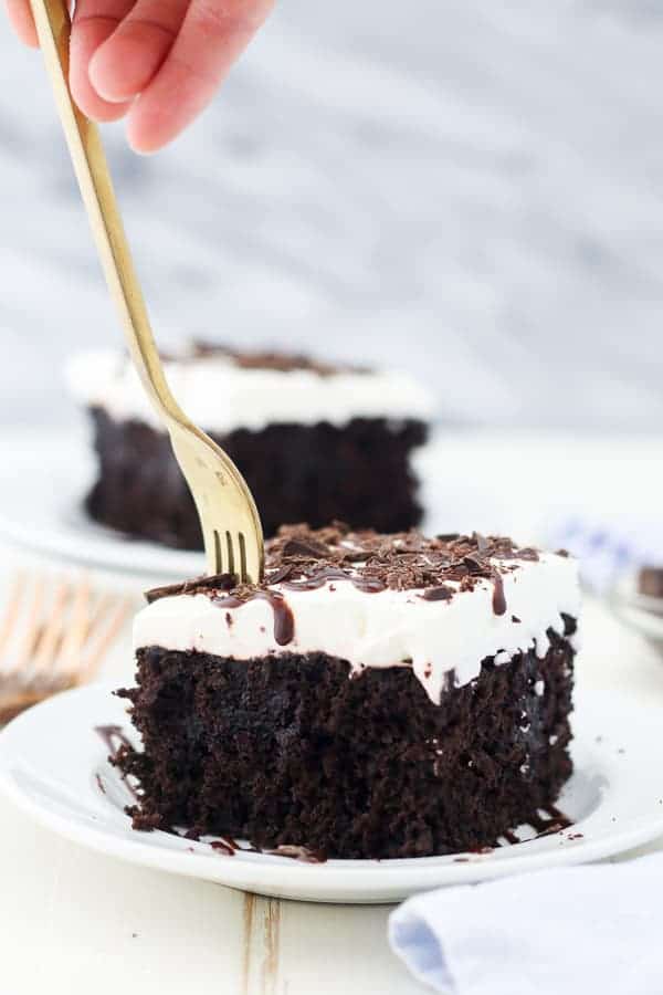 A gold fork stuck into a slice of chocolate poke cake garnished with whipped cream on a plate.