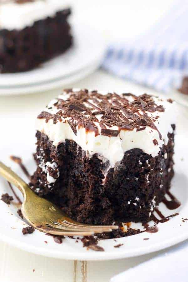 A slice of homemade chocolate poke cake on a plate, drizzled with chocolate sauce next to a gold fork.