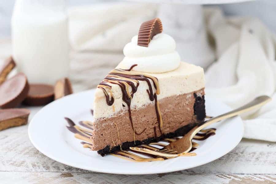 A gorgeous double layer peanut butter pie on a white plate with a gold fork. The pie is garnished with whipped cream, drizzles of peanut butter and chocolate and topped with a peanut butter cup.