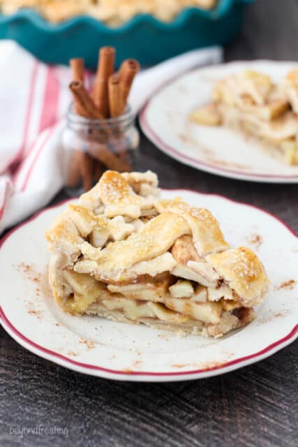 A large slice of apple pie with cinnamon sticks and a second slice in the background