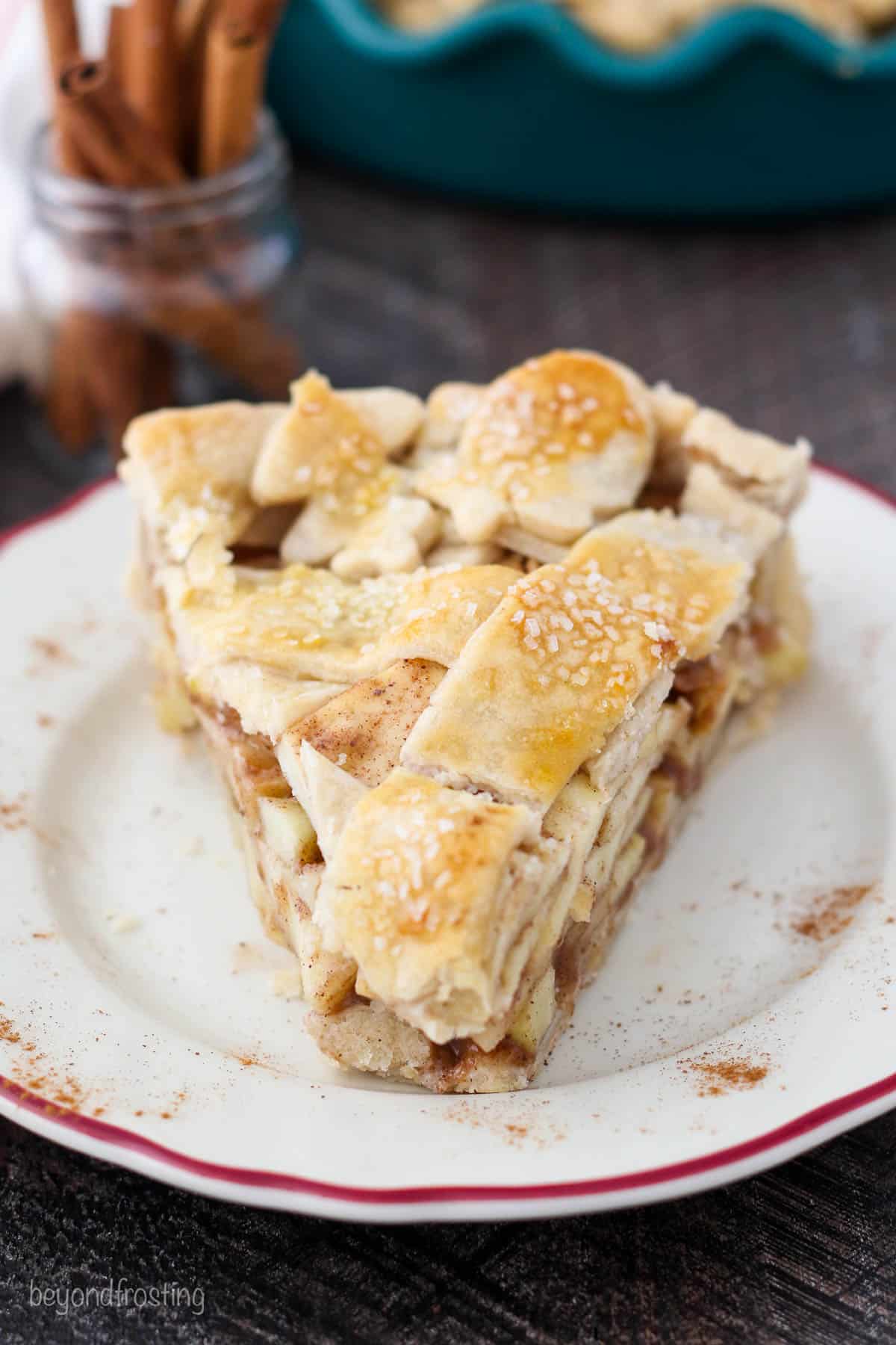 A head-on shot of a slice of apple pie with a jar of cinnamon sticks behind it