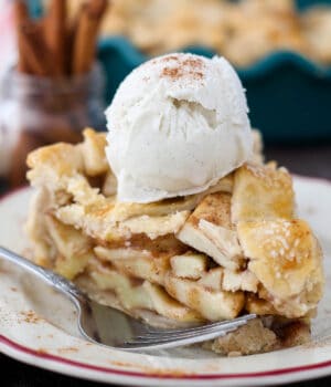 One slice of apple pie on a plate with a pie dish containing more slices in the background