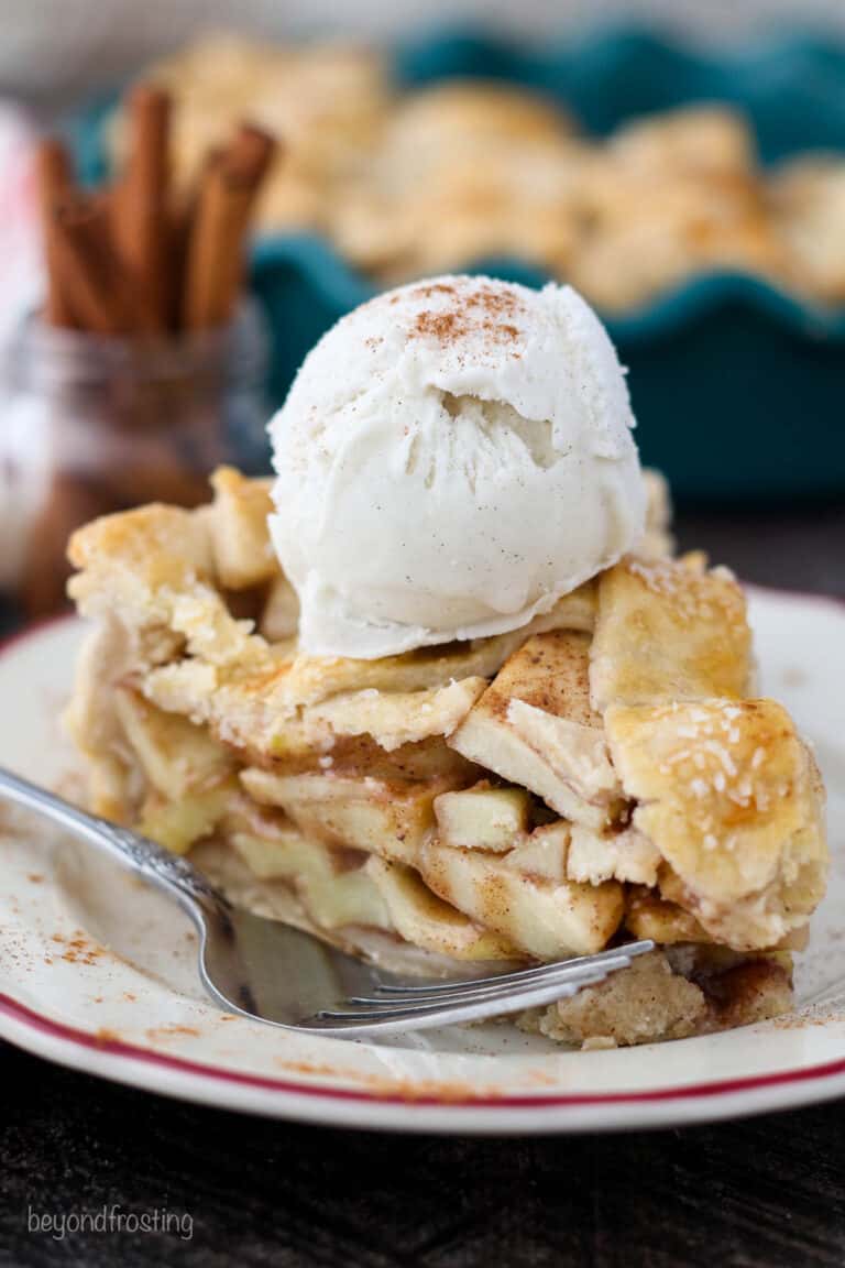 One slice of apple pie on a plate with a pie dish containing more slices in the background